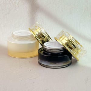 以下が含まれることがあります： 蓋付きの化粧品ジャーが3つ。1つのジャーは薄い黄色で、もう1つは黒です。蓋は透明で金色のアクセントが付いています。ジャーは明るい色の表面に置かれています。
