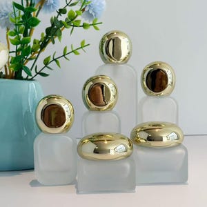 Op de afbeelding: Een verzameling flessen van matglas met goudkleurige, afgeronde deksels. De deksels hebben een reflecterend oppervlak. De flessen zijn gerangschikt op een wit oppervlak, met een vaas met bloemen op de achtergrond.