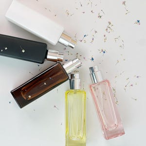 Könnte beinhalten: Draufsicht auf fünf rechteckige Parfümflaschen in Weiß, Schwarz, Braun, Gelb und Rosa. Jede Flasche hat einen silbernen Sprühkopf. Kleine getrocknete Blumen sind auf einer weißen Oberfläche um die Flaschen herum verstreut.