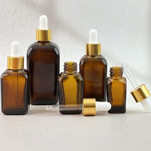 Puede incluir: Un surtido de frascos cuentagotas de vidrio ámbar con collares dorados y cuentagotas blancos. Las botellas varían en tamaño y forma, dispuestas sobre una superficie de color claro. Algunas botellas tienen cuentagotas insertados, otras están separadas.