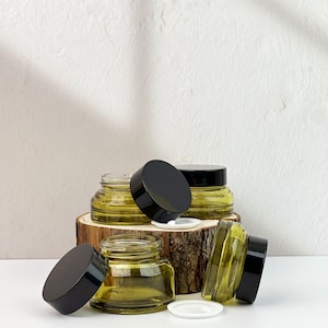 May include: A collection of small, clear glass jars with black lids, some open, are displayed on a wooden base. The jars have a subtle green hue. The background is a plain white wall. The jars are likely for storing small items.