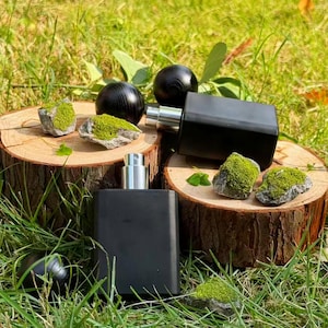Op de afbeelding: Twee zwarte rechthoekige parfumflessen met zilveren verstuivers worden gepresenteerd op houten boomstammen. Kleine rotsen bedekt met groen mos en twee zwarte houten bollen zijn ook aanwezig, tegen een achtergrond van groen gras.