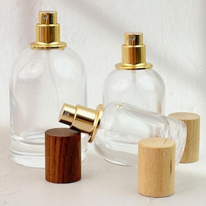 Op de afbeelding: Transparante glazen parfumflacons met goudkleurige sproeikoppen en houten doppen. De flacons hebben een afgeronde vorm. Eén fles ligt op zijn kant. Twee houten doppen zijn lichtgekleurd en één is donker.