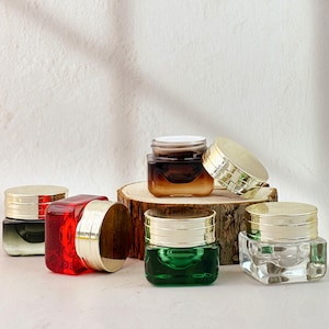 Könnte beinhalten: Eine Auswahl kleiner, quadratischer Gläser mit goldfarbenen Deckeln. Die Gläser sind mit Flüssigkeiten in verschiedenen Farben gefüllt, darunter Rot, Grün und Braun. Einige Gläser sind offen, andere geschlossen. Die Gläser sind auf einer Holzoberfläche angeordnet.