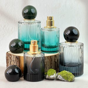 May include: Several glass perfume bottles with gold sprayers and decorative spherical stoppers. The bottles are various shades of blue and gray, with ribbed detailing. The bottles are arranged on a wooden surface with small rocks and moss.