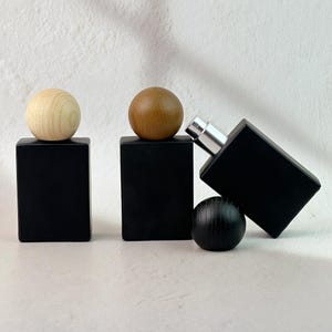 May include: Black rectangular perfume bottles with wooden spherical stoppers in various shades of brown and a silver atomizer. The bottles are arranged on a white surface, creating a minimalist aesthetic.