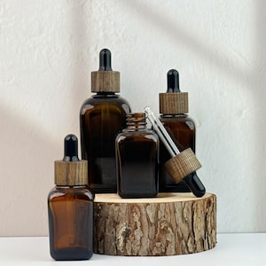 Könnte beinhalten: Eine Anordnung von braunen Glasflaschen mit schwarzen Pipetten und Holzkappen. Die Flaschen stehen auf einer Holzscheibe. Eine Flasche ist offen, mit einer Glaspipette. Der Hintergrund ist eine weiße Wand.