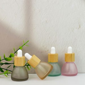 May include: Four small, frosted glass dropper bottles in shades of gray, peach, teal, and pink. Each bottle has a bamboo collar and a white dropper. The bottles are arranged on a white surface with green foliage in the background.