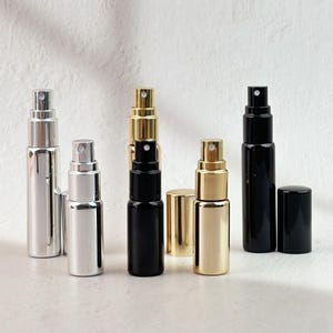 Op de afbeelding: Een assortiment kleine, navulbare parfum- of colognesprayflacons in zilver, goud en zwart. De flacons zijn cilindrisch met een sproeikop en een bijpassende dop. De afbeelding is gemaakt op een witte achtergrond.