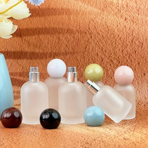 May include: A selection of frosted glass perfume bottles with silver atomisers and spherical caps in various colours. Several decorative spheres in brown, black, and blue are in front of the bottles. The background is a textured, warm-toned surface.