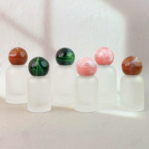 May include: A collection of frosted glass bottles, each topped with a unique, colorful gemstone sphere. The gemstones vary in color, including shades of red, green, and pink. The bottles are arranged against a white background.