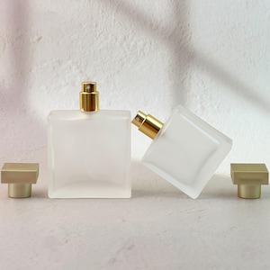 Puede incluir: Dos frascos de perfume de vidrio esmerilado con pulverizadores dorados y tapas cuadradas a juego. Los frascos son rectangulares y se apoyan sobre una superficie de color claro. Un frasco está en posición vertical y el otro está inclinado. El fondo es una pared blanca texturizada.