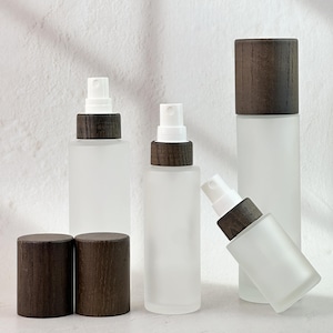 May include: A collection of frosted glass spray bottles with dark wood-effect caps and white spray nozzles. Two spare caps are in the foreground. The bottles are cylindrical and rectangular, suggesting a minimalist design for essential oils or perfumes.