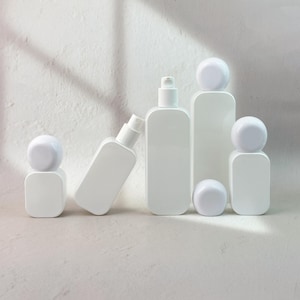 May include: A collection of five white cosmetic bottles with rounded rectangular bodies and spherical caps. Two bottles feature pump dispensers, while the others are capped. The containers are smooth, opaque, and arranged in a cluster.