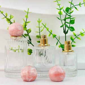 May include: Clear glass perfume bottles with gold-colored spray tops and pink marble-like stoppers. The bottles have a ribbed design and are arranged with green leafy sprigs. The stoppers are round and have a pink and white marbled appearance.