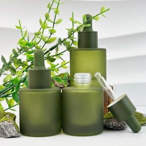 May include: Three olive green glass dropper bottles of varying sizes, with matching caps and droppers. One bottle is open, revealing the threads. The bottles are arranged with greenery and moss in the background, suggesting a natural skincare product.