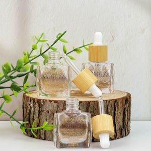 May include: Clear glass dropper bottles with gold geometric designs and bamboo lids are displayed on a wooden slice. The bottles are square-shaped and have a dropper with a white tip. Green leaves are in the background.