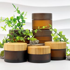 May include: Several amber-colored glass jars with wooden lids are arranged on a white surface. The jars vary in size and are stacked in some areas. Green leafy sprigs and small rocks with moss are placed around the jars, creating a natural aesthetic.
