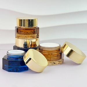 Op de afbeelding: Een verzameling cosmetische potten in verschillende maten en kleuren. De potten zijn gemaakt van glas met goudkleurige deksels. Sommige potten zijn amberkleurig, andere blauw. De potten zijn op een wit oppervlak geplaatst.