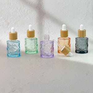 Puede incluir: Cinco frascos cuentagotas de vidrio pequeños con patrones geométricos facetados. Cuatro frascos tienen collares de bambú y bulbos de goma blancos, mientras que un frasco morado está abierto. Los colores incluyen azul claro, verde claro, morado, naranja y gris oscuro.