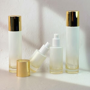 May include: Several clear glass cosmetic bottles with gold-colored lids and white pumps. The bottles have a gradient effect, transitioning from white to a pale yellow. The bottles are cylindrical and of varying heights.