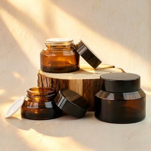 May include: Three amber glass cosmetic jars with wide openings and black screw-on lids. Two jars are open with white plastic inner liners visible, while one jar is closed with its black lid. The jars have a smooth, rounded base and a thick glass wall.