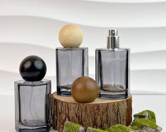 May include: Three gray glass perfume bottles with wooden and silver spray tops. One bottle has a black wooden sphere, another a light wood sphere, and the third a silver spray top. The bottles are displayed on a wood slice with mossy rocks.