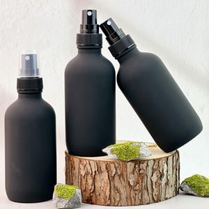 Puede incluir: Tres botellas de spray negras mate con boquillas de spray negras. Una botella tiene una tapa de plástico transparente. Las botellas están dispuestas sobre una rodaja de madera con pequeñas rocas cubiertas de musgo verde.