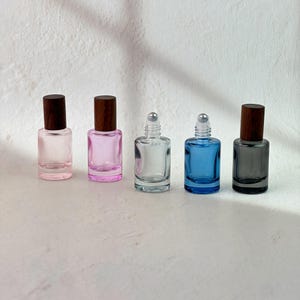 May include: Five small glass roller bottles arranged in a row. The bottles feature cylindrical bodies in light pink, pink, clear, blue, and dark gray. Each bottle is topped with a dark brown wooden cap and a silver metal roller ball applicator.