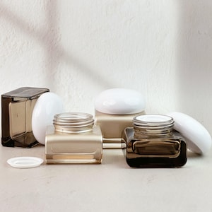 Op de afbeelding: Een assortiment glazen potten en containers in verschillende vormen en maten. De potten zijn in bruin- en goudtinten, met witte deksels. De containers zijn gerangschikt op een lichtgekleurd oppervlak, wat een minimalistische esthetiek creëert.