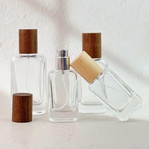 May include: Clear glass perfume bottles with wooden caps. One bottle has a silver atomizer. The bottles are rectangular with rounded corners. The wooden caps are cylindrical and a medium brown color. The bottles are empty.