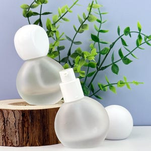 May include: Two frosted glass bottles with white tops and a white sphere are displayed. The bottles have a rounded shape and sit on a wooden base. Green leafy branches are in the background, with a light blue wall.