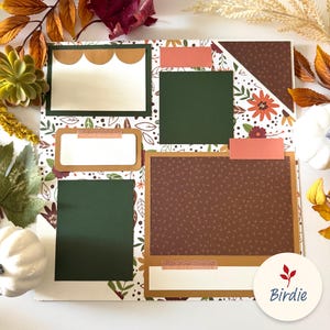 May include: A scrapbook page with a fall theme, featuring a patterned background with leaves and floral designs. It includes photo mats in green, brown, and white, accented with decorative tape. The page is adorned with fall foliage and a white pumpkin.