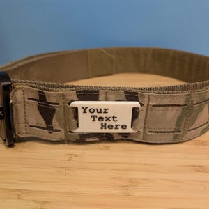 May include: Camouflage-patterned belt with a black buckle and a white rectangular tag that reads "Your Text Here". The belt is made of durable fabric and is designed for functionality.