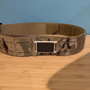 May include: A tactical belt with a camouflage pattern. The belt features a black and white rectangular buckle and is made of durable, woven material. The belt is designed for secure and comfortable wear.
