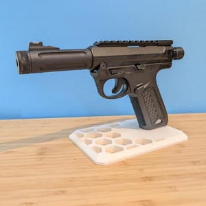 May include: A black handgun with a cylindrical barrel and a textured grip, displayed on a white honeycomb-patterned stand. The gun features a top rail and a sight. The background is a light blue wall and a wooden surface.