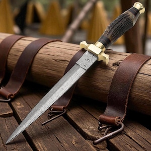 Puede incluir: Primer plano de una daga con una hoja estampada y un mango marrón oscuro. La daga tiene una guarda y pomo dorados. Descansa sobre una superficie de madera con correas de cuero.