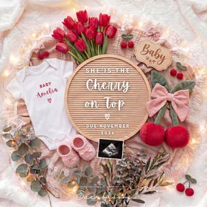 May include: A white baby bodysuit with the text Baby Amelia and a heart, a round letter board reading She is the Cherry on Top Due November 2026, pink knit booties, red tulips, a sonogram, a pink bow, and plush red cherries on a textured fabric.