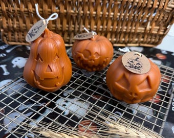 May include: Three orange pumpkin-shaped candles with carved jack-o'-lantern faces. Each candle has a small tag attached with a string. The candles are on a metal cooling rack, with a wicker basket in the background. The tag on the right candle says "Pumpkin Pie".