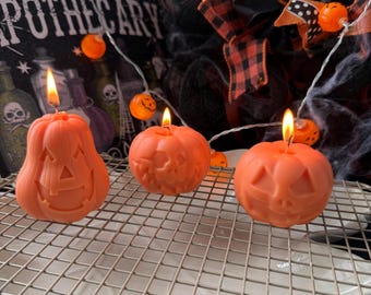 May include: Three orange pumpkin-shaped candles with lit wicks sit on a metal rack. The candles have carved jack-o'-lantern faces. A string of small orange lights and a Halloween wreath are in the background. The word "APOTHECARY" is visible.