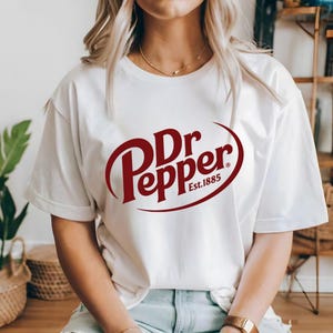 Puede incluir: Camiseta blanca con el logotipo de Dr Pepper en color granate. El logotipo incluye las palabras "Dr Pepper" en una fuente estilizada, con "Est. 1885" debajo. La camiseta es de cuello redondo.