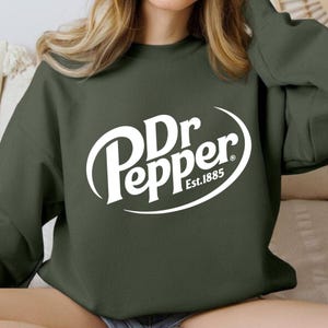 Puede incluir: Una sudadera verde oscuro con el logo de Dr Pepper en blanco. El logo incluye el nombre de la marca y "Est. 1885" en una fuente estilizada. La sudadera es de color liso y tiene cuello redondo.
