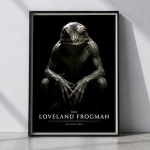 May include: A framed vertical poster featuring a dark, textured illustration of a humanoid frog creature in a crouching position against a black background. The text reads THE LOVELAND FROGMAN and Loveland, Ohio in white serif lettering.