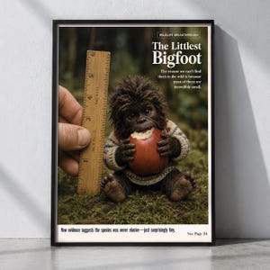 May include: A framed poster titled The Littlest Bigfoot featuring a small, furry, dark-brown creature wearing a patterned sweater and eating a red apple. A wooden ruler shows the creature is approximately 6 inches tall. Text includes Wildlife Breakthrough.