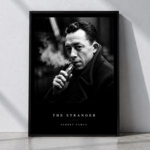 May include: A black and white framed poster featuring a portrait of Albert Camus smoking, with the text THE STRANGER and ALBERT CAMUS printed in white at the bottom. The image shows a man in a dark coat with smoke rising from a handheld device.