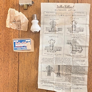 May include: A vintage cupboard latch set with a white ceramic knob, metal hardware, and an instruction sheet. The set includes a small box with the words "Mother Hubbard Cupboard Latch" printed on it. The instruction sheet provides diagrams and instructions.