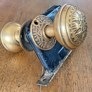May include: Antique brass door knobs with a black metal plate. The knobs have a detailed floral design and are likely from a vintage door set. The hardware is aged with signs of wear.