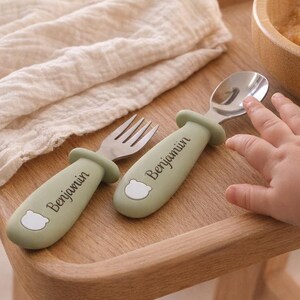 May include: A baby fork and spoon set with green handles and the name "Benjamin" printed on them. The utensils are made of stainless steel and have a bear design. The set is on a wooden table next to a bowl of baby food.
