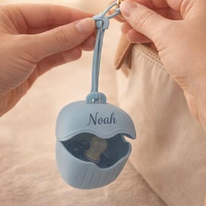 May include: A light blue pacifier case with the name "Noah" printed on the front. The case is open, revealing a pacifier inside. The case is attached to a light blue strap and a gold-colored clasp.
