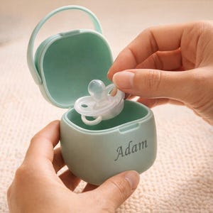 May include: A light green pacifier case with a handle, holding a clear pacifier. The case is open, and the name "Adam" is printed on the front. The pacifier is being held by a person.